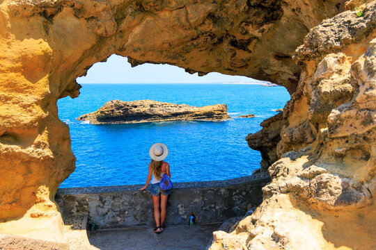 Rear view of female tourist with straw hat looking through rock arch frame at ocean in Biarritz France