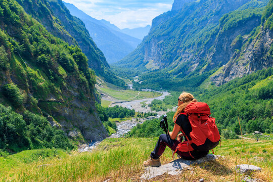 Female hiker with red backpack sitting on rock looking down at green mountain valley in Sixt Fer a Cheval, Alps in France, Chamonix