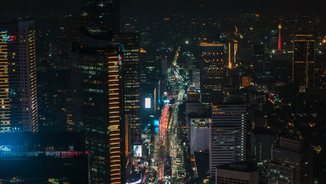Aerial view of the illuminated Jakarta skyline at night featuring modern skyscrapers, busy traffic on city streets, and the distant glowing National Monument in Jakarta, Indonesia.