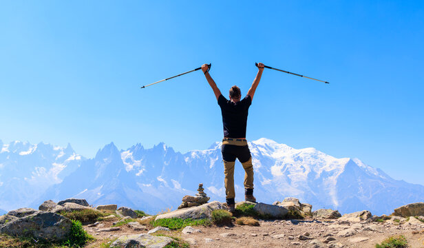 Rear view of male hiker raising trekking poles in success on mountain peak looking at Mont Blanc, Alps, France