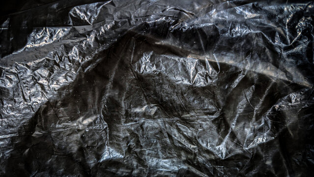 Dark crumpled plastic sheet background with strong highlights and shadows creating a deep textured surface for design.