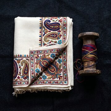 Traditional Indian Pashmina Shawl & Spool: A neatly folded, authentic cashmere Pashmina shawl with colorful paisley (Kalka) embroidery, resting next to a rustic wooden thread spool,