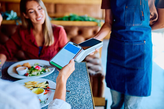 Customer paying restaurant bill with mobile phone, holding it over a payment terminal held by a server, demonstrating modern banking technology