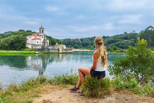 Rear view of female tourist sitting on coast looking at Church of Our Lady of Sorrows in Niembro, Spain