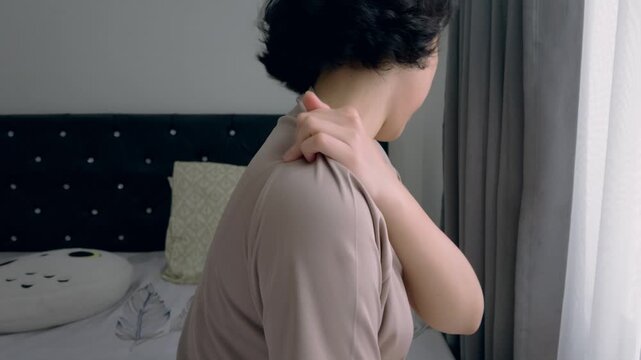 A woman sits on a bed near a window rubbing her shoulder and neck, showing stiffness and discomfort.