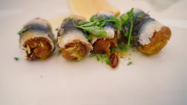 Golden stuffed sardines rolled with breadcrumb, herbs and citrus rest on a white plate, close up.