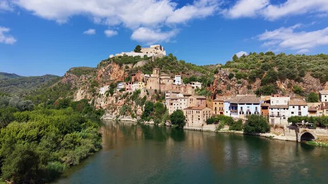 Miravet village perched on rocky cliffs above the Ebro River in Catalonia, Spain, with a medieval Templar castle crowning the hilltop under blue summer skies