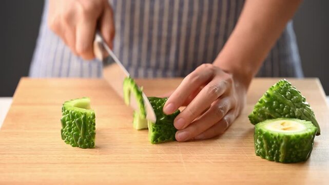 Woman hands holding a kitchen knife and slicing bitter gourd on a wooden cutting board