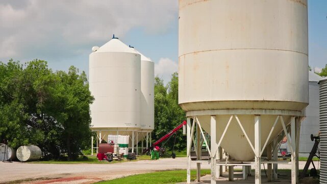 Static wide shot of white grain silos and farm equipment on a prairie farm with trees swaying beneath a bright blue summer sky