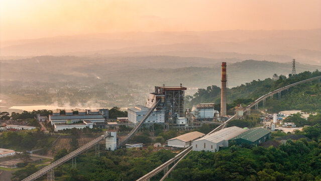 Aerial view of a coal-fired power plant with a tall chimney, conveyor belts, and industrial buildings nestled in a hilly, forested landscape during the golden hour in Indonesia.