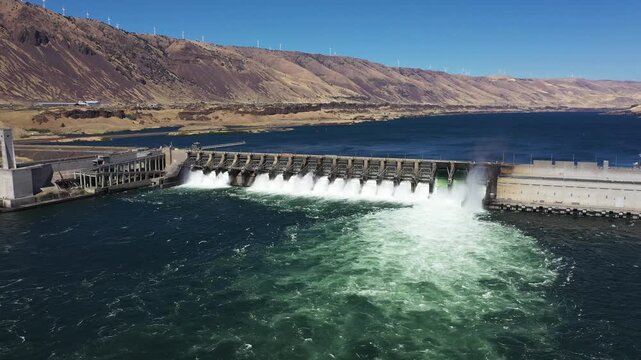 4K aerial drone footage of John Day Dam near Rufus, Oregon, USA with distant wind turbines. Hydroelectric power station, renewable energy, pelicans fishing in Columbia River Gorge