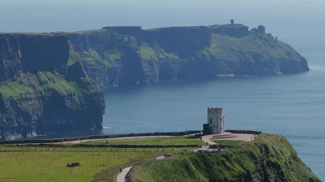 People follow a coastal path around OBrien Castle on a grassy headland with the tall Cliffs of Moher dropping to the calm Atlantic below.