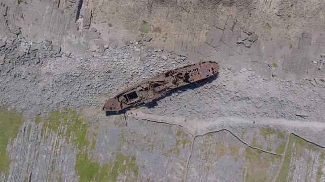Rusting Plassey cargo ship lies stranded on a bare rock shoreline as the camera hovers directly overhead in bright daylight.