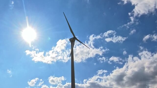 powerful wind turbine stands tall against brilliant blue sky with bright sun rays shining intensely through scattered white clouds, symbolizing clean energy and renewable power sources for