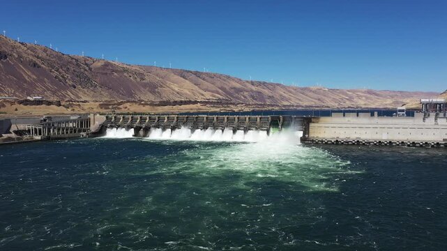 4K aerial drone footage of John Day Dam near Rufus, Oregon, USA with distant wind turbines. Hydroelectric power station, renewable energy, pelicans fishing in Columbia River Gorge