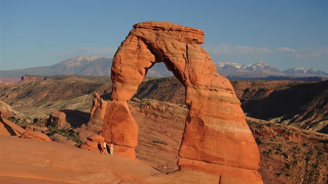 Iconic Delicate Arch stands against blue sky with snow-capped La Sal Mountains in background at Arches National Park Utah. Hikers explore red sandstone formation during golden hour light.