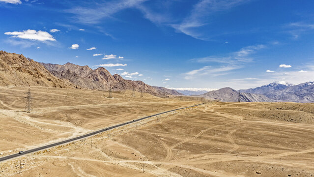 Aerial view of a long road winding through the arid, mountainous landscape of the Himalayas under a clear blue sky in Leh, Ladakh, India.