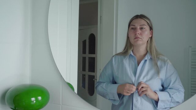 Job applicant buttons a light work shirt in front of a mirror, steadying herself before a remote job interview.