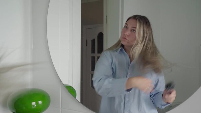 Job applicant in an oversized button-up calmly brushes her long hair in front of a round bathroom mirror before a video call.