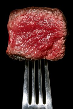 Close up of rare medium cooked beef steak cube impaled on metal fork against plain solid black background showing juicy red meat texture