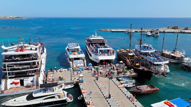 Aerial view inside Paphos Harbour in Cyprus. Fishing boats, tour boats, marina piers and clear blue water create a colourful Mediterranean waterfront view.