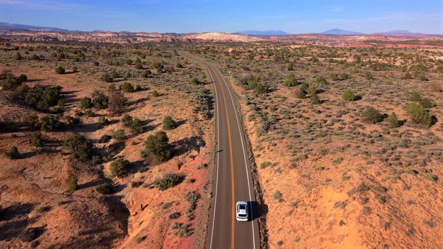 Aerial drone footage of Utah Scenic Byway 12 showing white car traveling along winding desert road surrounded by red rock terrain and scattered juniper trees with colorful cliffs on horizon.