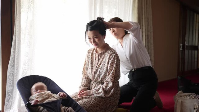 Soft light bathes a cozy, warm room. Two women smile, one gently styling the other's hair. Baby rests in a bassinet nearby, calm and secure. This intimate family moment conveys quiet care