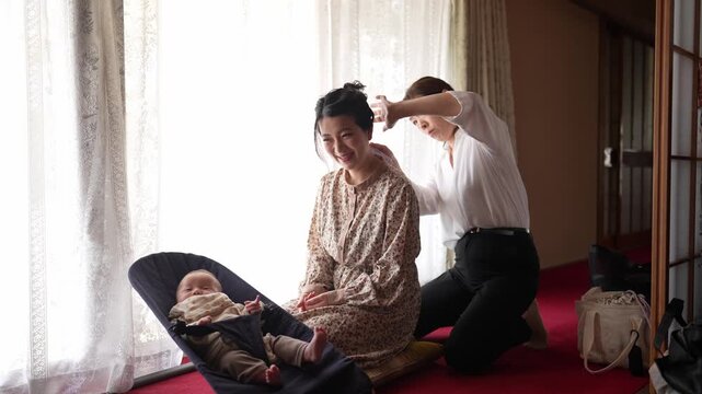 Soft light bathes a quiet Japanese room. Two women kneel, one gently combing the other's hair. Baby rests peacefully in a carrier nearby. Warmth radiates from their calm, focused expressions