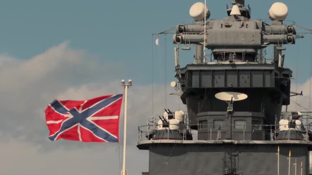 Close up telephoto view of Russian naval jack flag waving in wind on grey military cruiser warship superstructure with radar antennas and defense guns against blue sky with clouds during sunny day