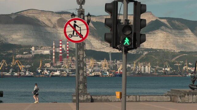 Pedestrian traffic light changing from red to green and no scooter sign at Novorossiysk sea port bay. Blurred cars pass by in foreground while people walk dogs against mountain summer background.