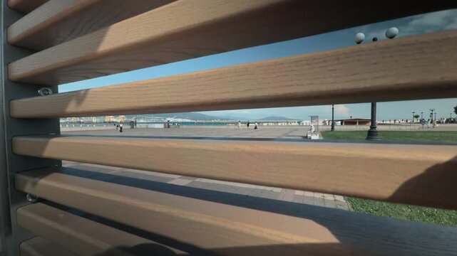 Looking through wooden bench slats at paved city embankment square with walking anonymous people green grass sea bay and distant mountains in Novorossiysk on a bright sunny summer day candid view