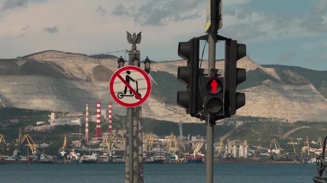 Red pedestrian traffic light and no scooter road sign against beautiful industrial seaport with yellow cranes ships and large mountains at Novorossiysk city bay coast during sunny summer daylight