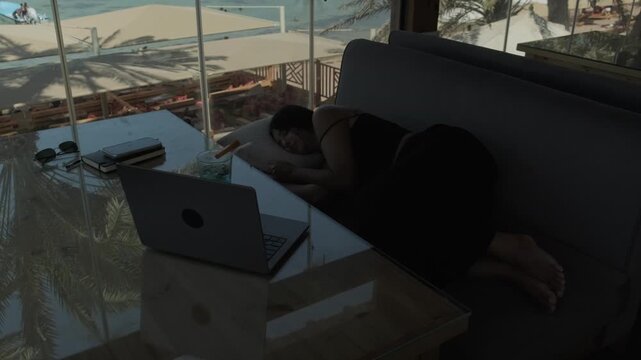 Female digital nomad choosing to relax instead of working, getting comfortable on a sofa beside her laptop in a tropical cafe with ocean views and palm trees