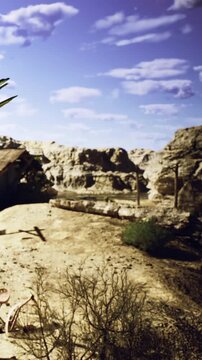 Beneath a vast blue sky adorned with clouds, rocky cliffs rise from the arid desert. Sparse shrubs dot the sandy terrain, echoing natures stunning, rugged beauty at dusk.