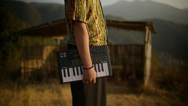 Young man in patterned shirt holding a MIDI keyboard standing beside a bamboo hut overlooking layered mountain ranges at golden hour