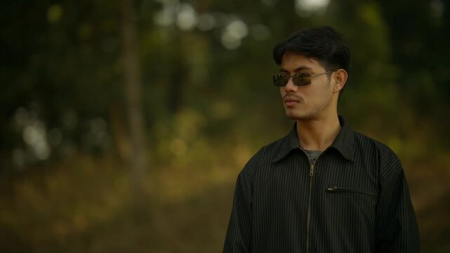 Close portrait of a young man in a striped black jacket and sunglasses holding a MIDI keyboard against a blurred forest backdrop at sunset