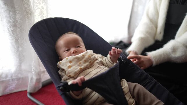 Baby sleeps peacefully in a dark blue bouncer. Soft light streams through lace curtains behind. Red carpet adds warmth to the cozy room. Parent's hands gently adjust the baby's position