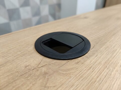 Black round cable management grommet installed in a light wood office desk surface for organized computer wire routing