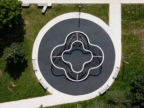 Aerial view of a modern climbing frame in a playground, surrounded by a circular rubber surface, green grass, and park benches in Kaunas, Kaunas County, Lithuania.