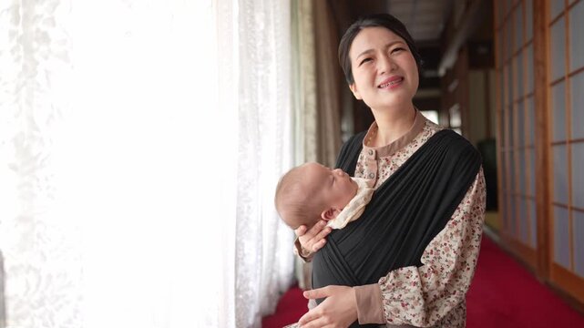 Mother smiles gently with baby in traditional wrap. Soft light spills through window beside her. Red carpet leads to wooden sliding doors. Warm, peaceful moment with child close