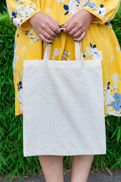 Woman Holding Blank Cotton Tote Bag in Yellow Floral Dress Against Green Foliage