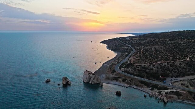 Wide aerial view of Petra tou Romiou at golden sunset in Cyprus. Sea stacks, curved coastline and calm Mediterranean water create a peaceful coastal panorama.