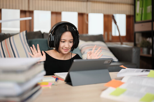 Asian teenage girl greeting in online class, remote learning at home using digital tablet, writing and reading textbooks, self-study for back to school, Homeschooling, university education lifestyle.
