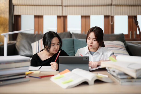 Two Asian teenage girls concentrate on online class, remote learning at home using tablet, writing and reading textbooks, self-study for back to school, Homeschooling, university education lifestyle.