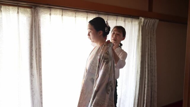 Soft light streams through lace curtains. Two women stand near a window. One gently helps the other don a kimono. Their expressions show quiet focus. Moment of grace, tradition, and care