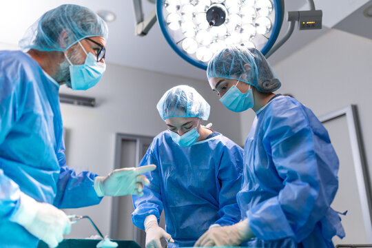 Experienced surgeon leading a medical team during an operation in a brightly lit hospital operating room. Doctors wearing surgical scrubs, masks, and caps concentrating on performing procedure