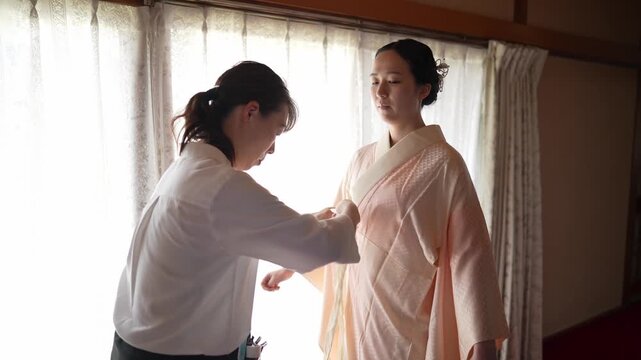 Soft light streams through sheer curtains. One woman adjusts the other's robe gently. They share quiet moments of care. Ideal for bridal or cultural scenes. No animals or plants present
