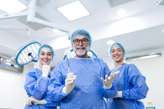 Confident senior surgeon holding surgical instruments while smiling with his female medical team in a bright modern hospital operating room. Doctors in blue scrubs and caps looking directly at camera