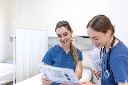 Two smiling young female doctors in blue medical scrubs reviewing patient chart documents and discussing medical treatment plans inside a bright hospital ward room environment.