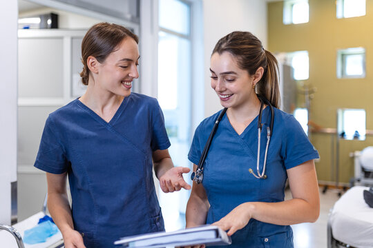 Two smiling young female doctors in blue scrubs discussing medical notes and patient documents while standing in a bright hospital corridor or ward room during their daily healthcare shift.